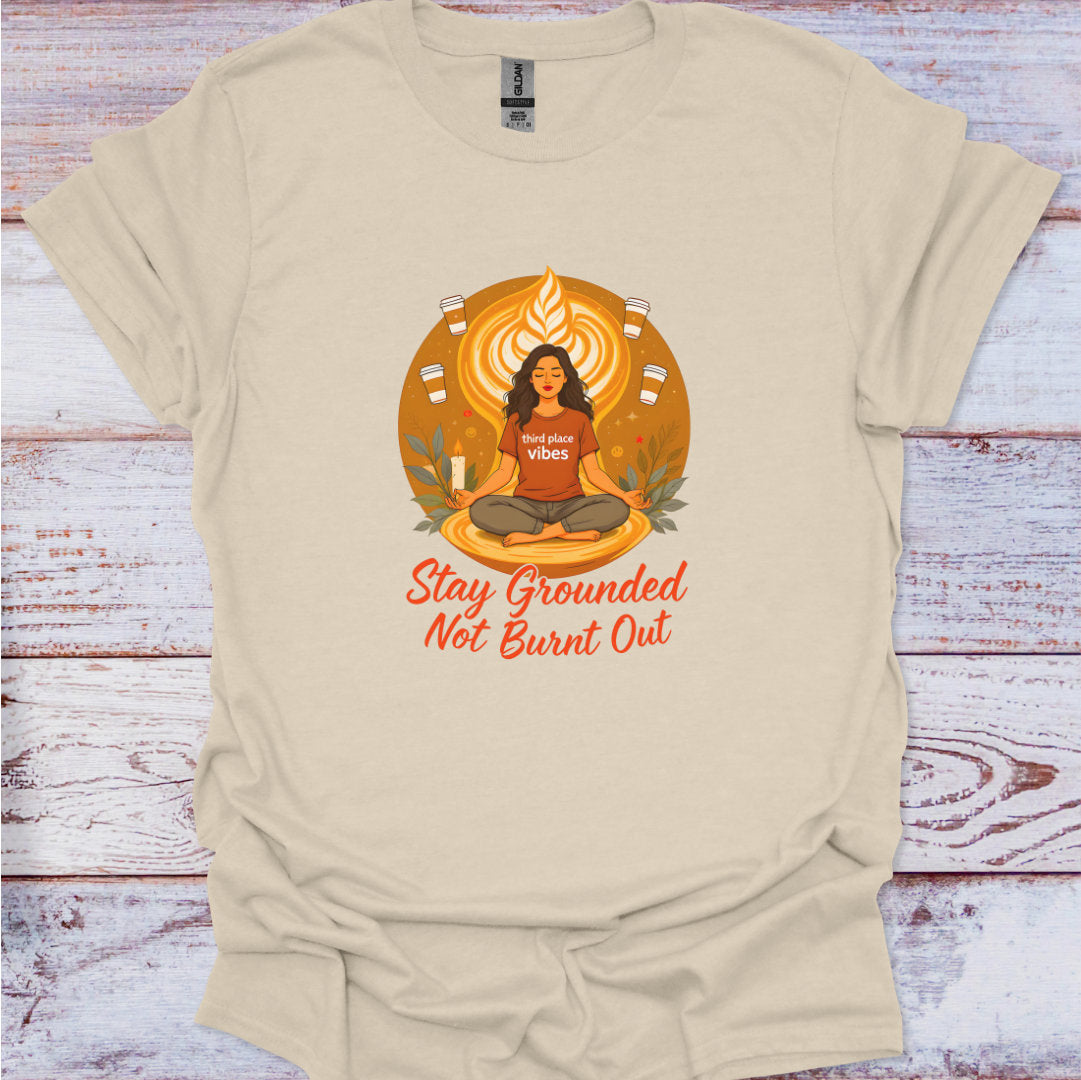 Beige t-shirt with a graphic of a woman in meditation and text 'Stay Grounded Not Burnt Out' on a wooden surface.
