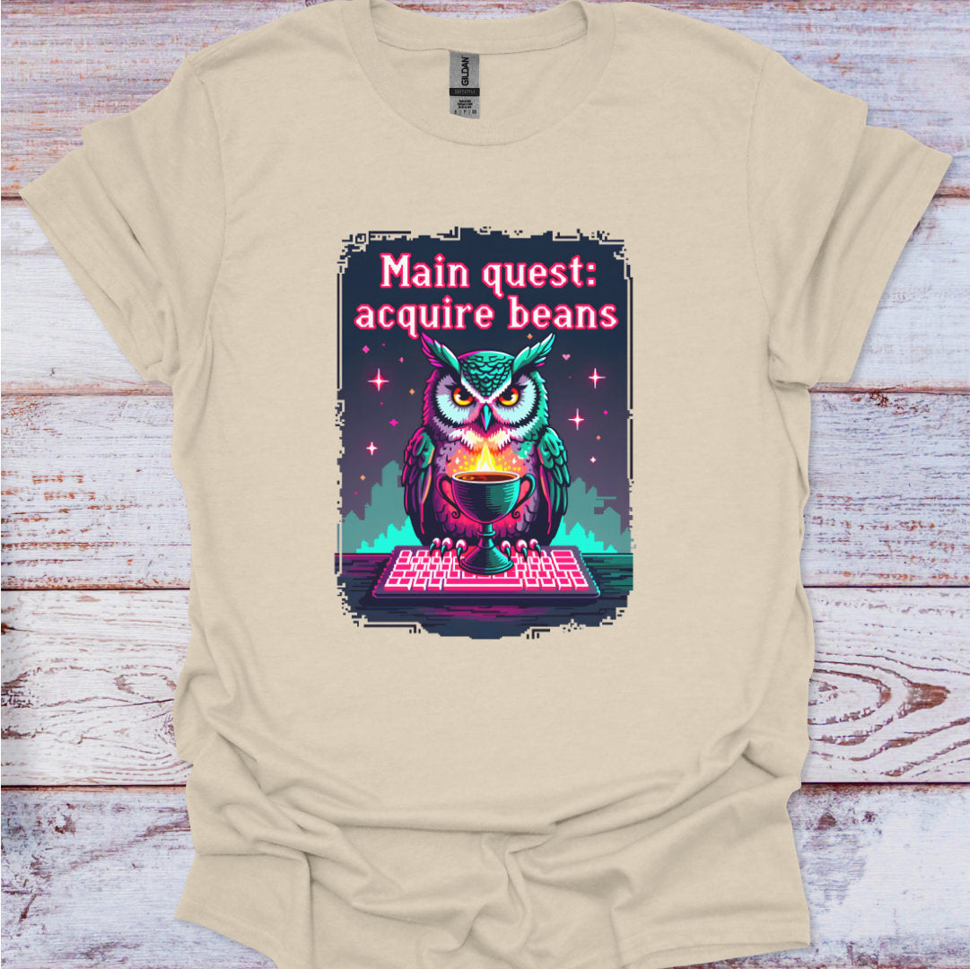 Beige t-shirt with an owl graphic and text 'Main quest: acquire beans' on a wooden surface.