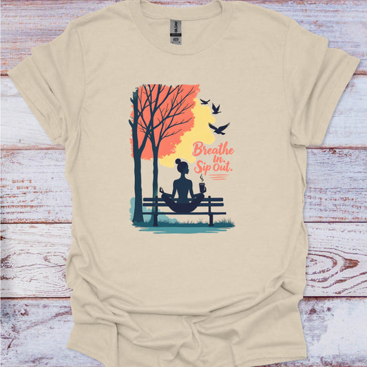 Beige t-shirt with a graphic of a woman meditating by a tree, birds, and text 'Breathe In Sip Out' on a wooden surface.