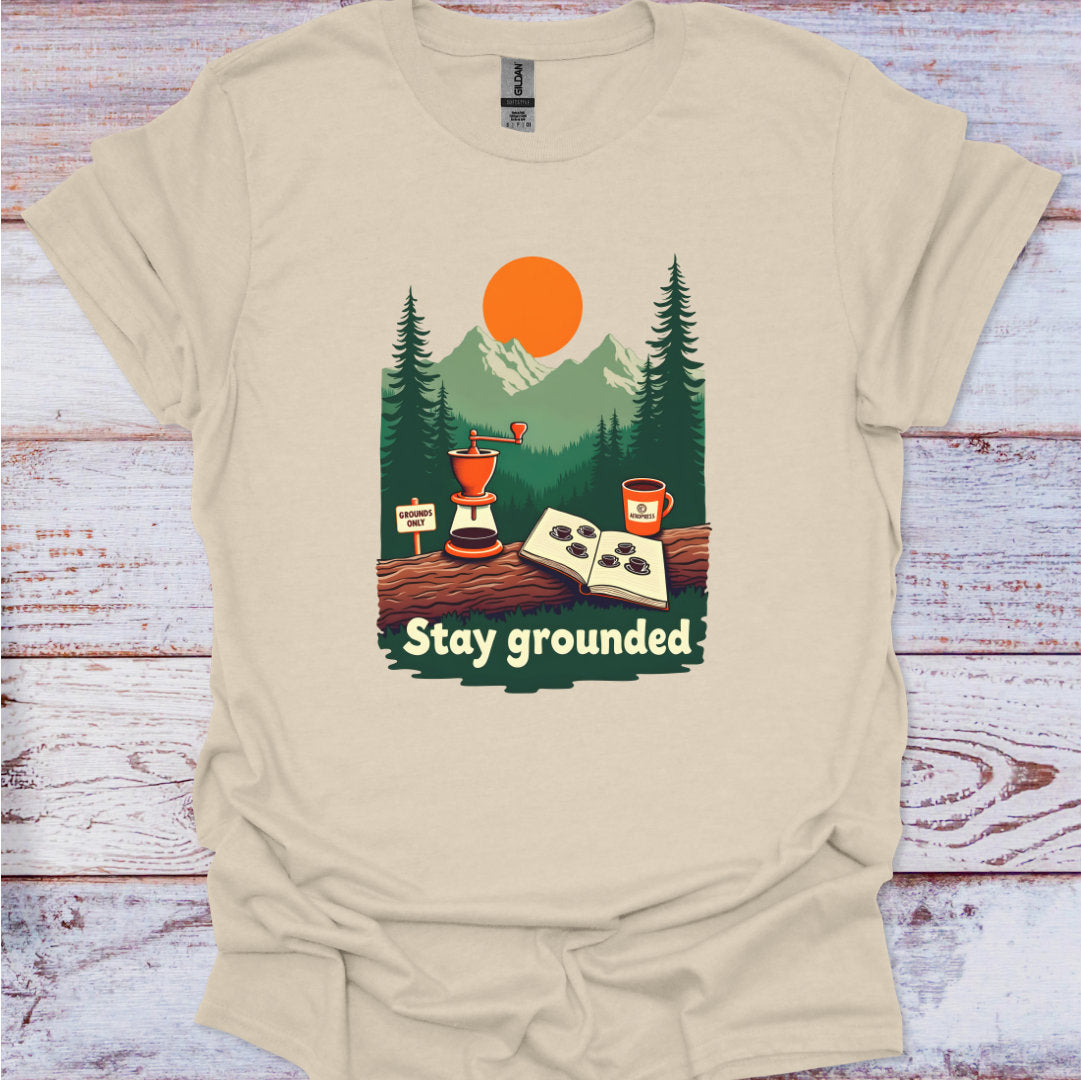 Beige t-shirt with a graphic design of a forest scene, coffee grinder, and cup, with 'Stay grounded' text, on a wooden surface.