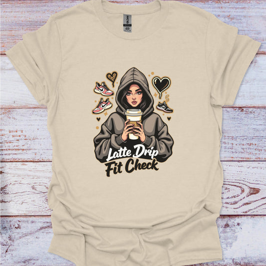 Beige t-shirt with graphic design of a person in a hoodie holding a coffee cup, text 'Latte Drip Fit Check', on a wooden surface.