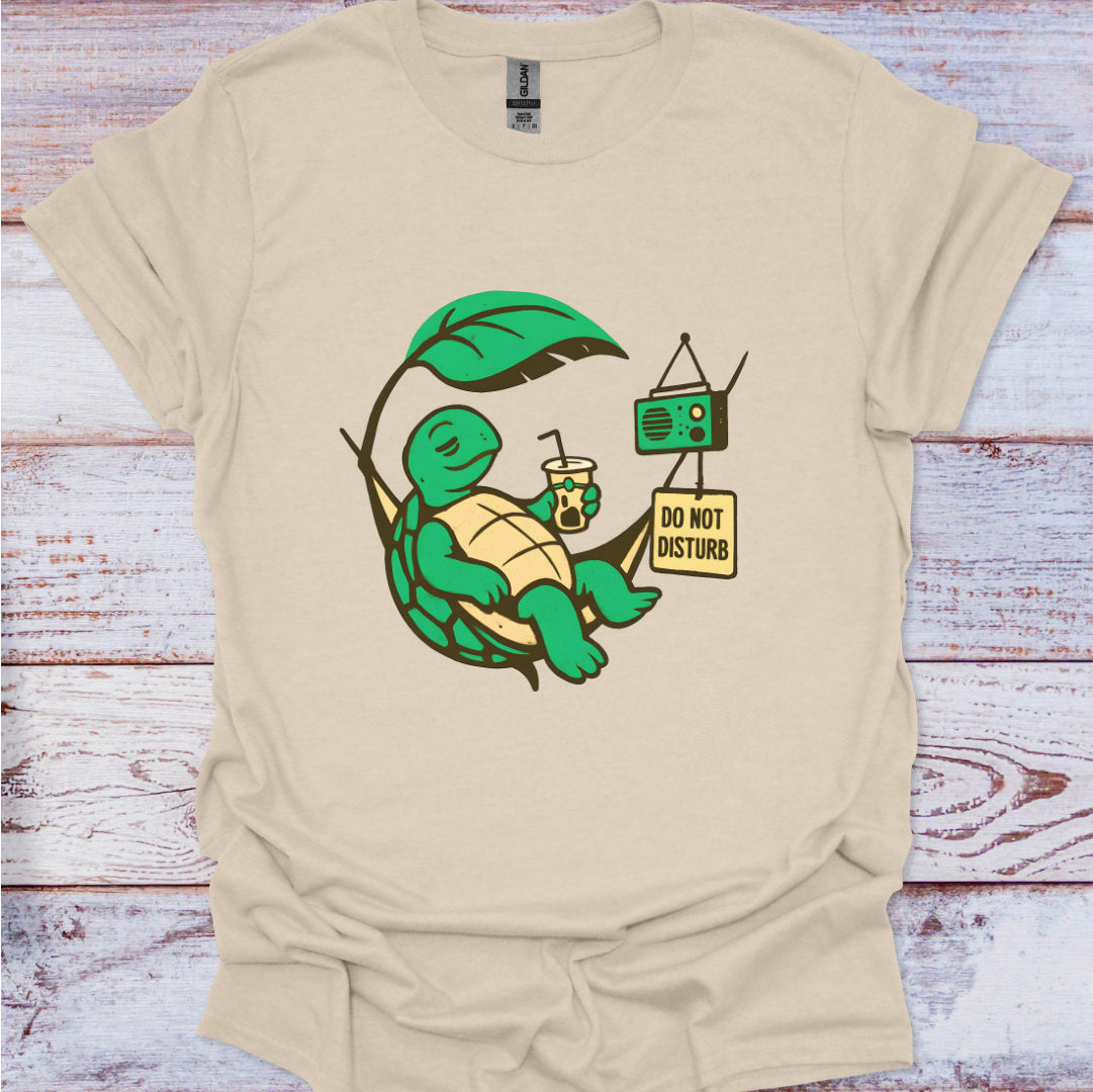 Beige t-shirt with a turtle graphic holding a leaf, drink, and radio on a wooden surface