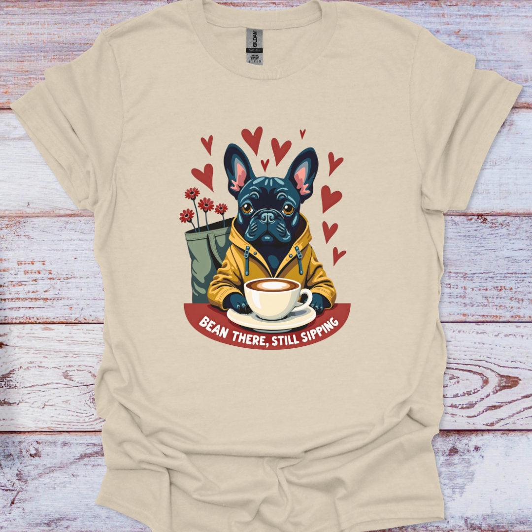 Cream t-shirt with a graphic of a dog holding a coffee cup and text 'Bean There, Still Sipping' on a wooden surface.