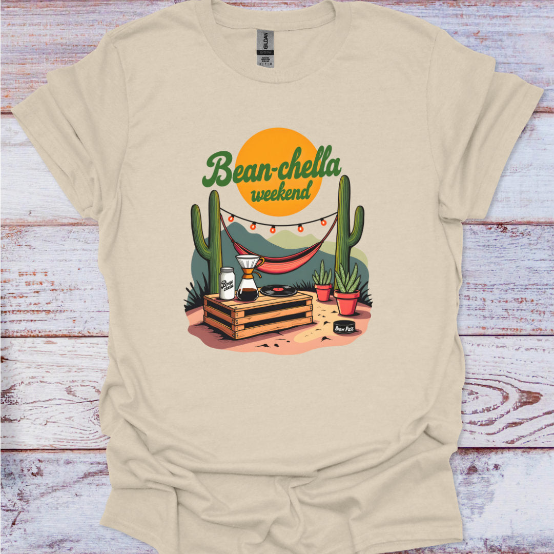 Beige t-shirt with a graphic design featuring cacti, a hammock, and 'Bean-chella weekend' text on a wooden surface.