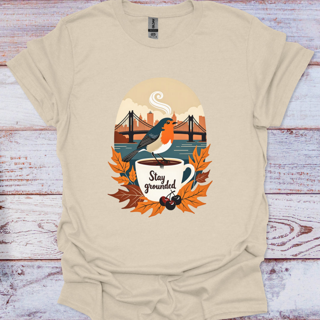 Beige t-shirt with a graphic design of a bird on a coffee cup with autumn leaves and text 'Stay grounded' on a wooden surface.