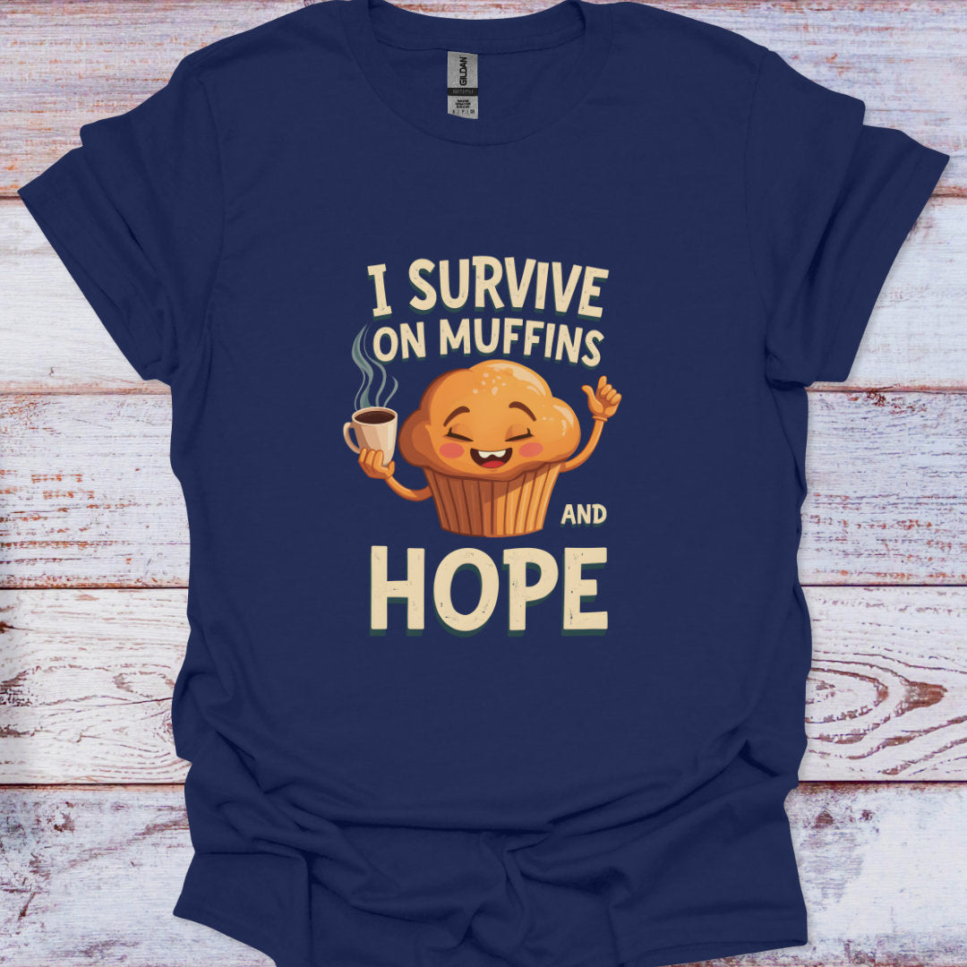 Navy blue t-shirt with a cartoon muffin holding a coffee cup and text 'I Survive on Muffins and Hope' on a wooden surface.