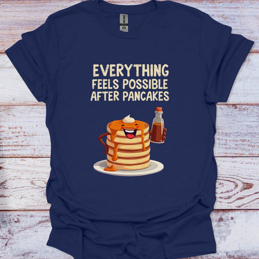 Navy blue t-shirt with a graphic of pancakes and syrup and text 'Everything feels possible after pancakes' on a wooden surface.
