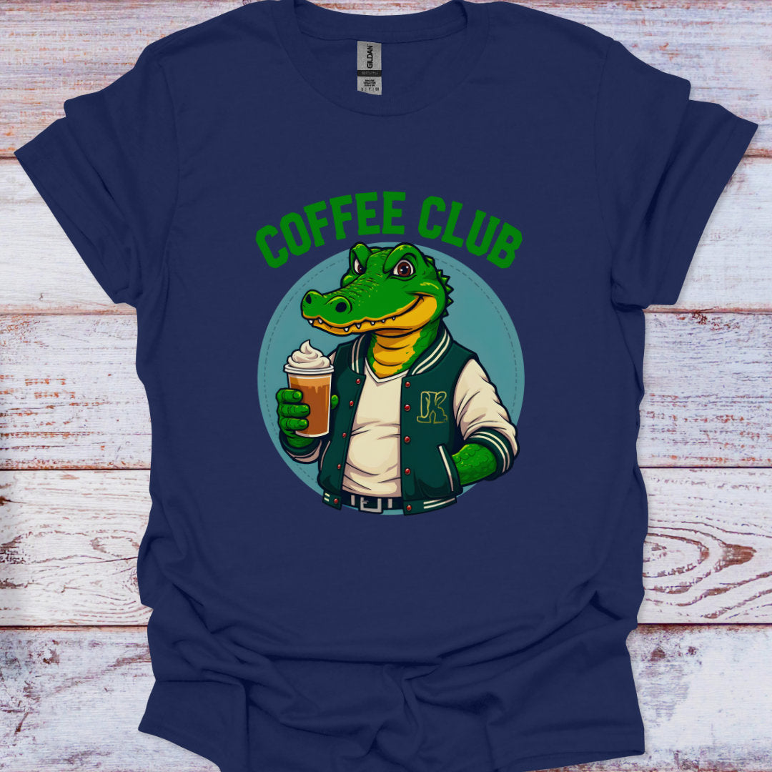 Navy blue t-shirt with a graphic of a crocodile in a coffee club jacket holding a coffee cup, on a wooden surface.