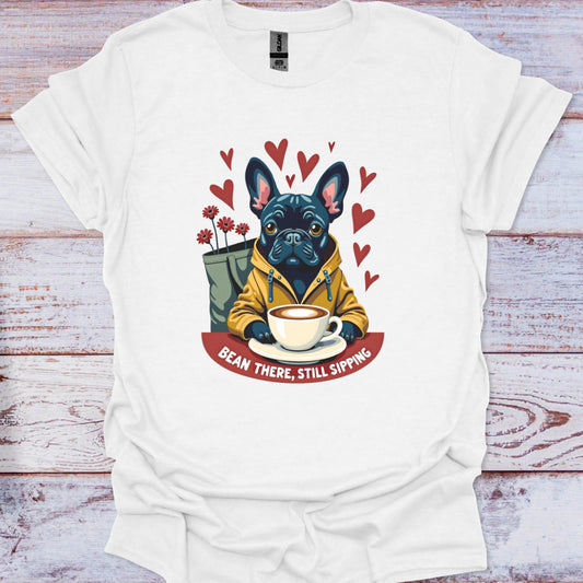 White t-shirt with a graphic of a dog holding a coffee cup and text 'Bean There, Still Sipping' on a wooden surface.