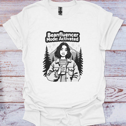 White t-shirt with a graphic of a person holding a coffee cup and text 'Beanfluencer Mode: Activated' on a wooden surface.