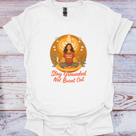 White t-shirt with a graphic design of a person in a meditative pose and text 'Stay Grounded Not Burnt Out' on a wooden surface.