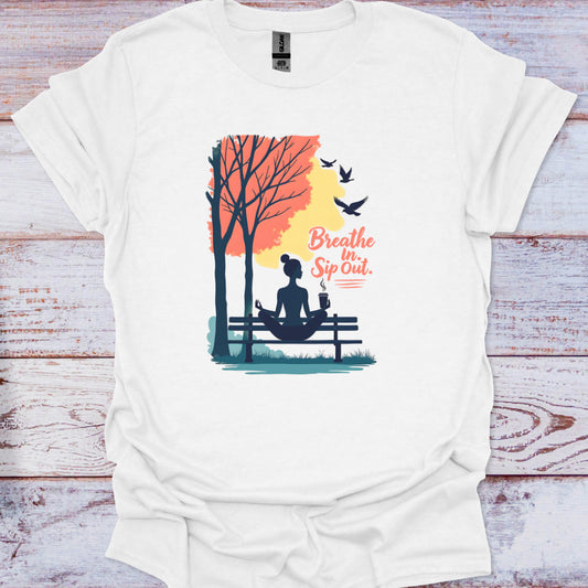 White t-shirt with a graphic of a woman sitting on a bench in a park, trees, and birds with text 'Breathe In Sip Out' on a wooden surface.
