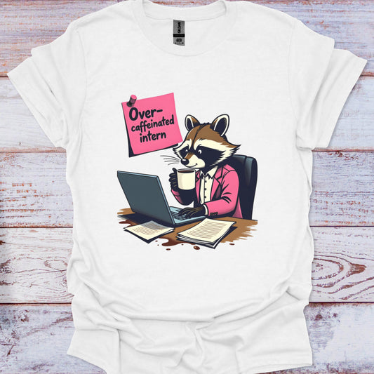 White t-shirt with a raccoon illustration holding a coffee cup and using a laptop, on a wooden surface.