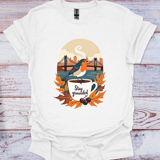 White t-shirt with a graphic design of a bird on a coffee cup with leaves and text 'Stay grounded' on a wooden background.