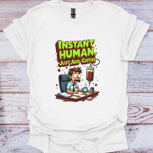 White t-shirt with a cartoon design and text 'Instant Human, Just Add Coffee' on a wooden surface.